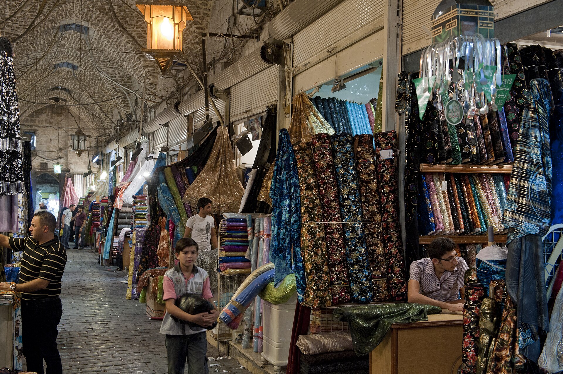 A covered market lane in Aleppo lined with stalls of printed and woven textiles.