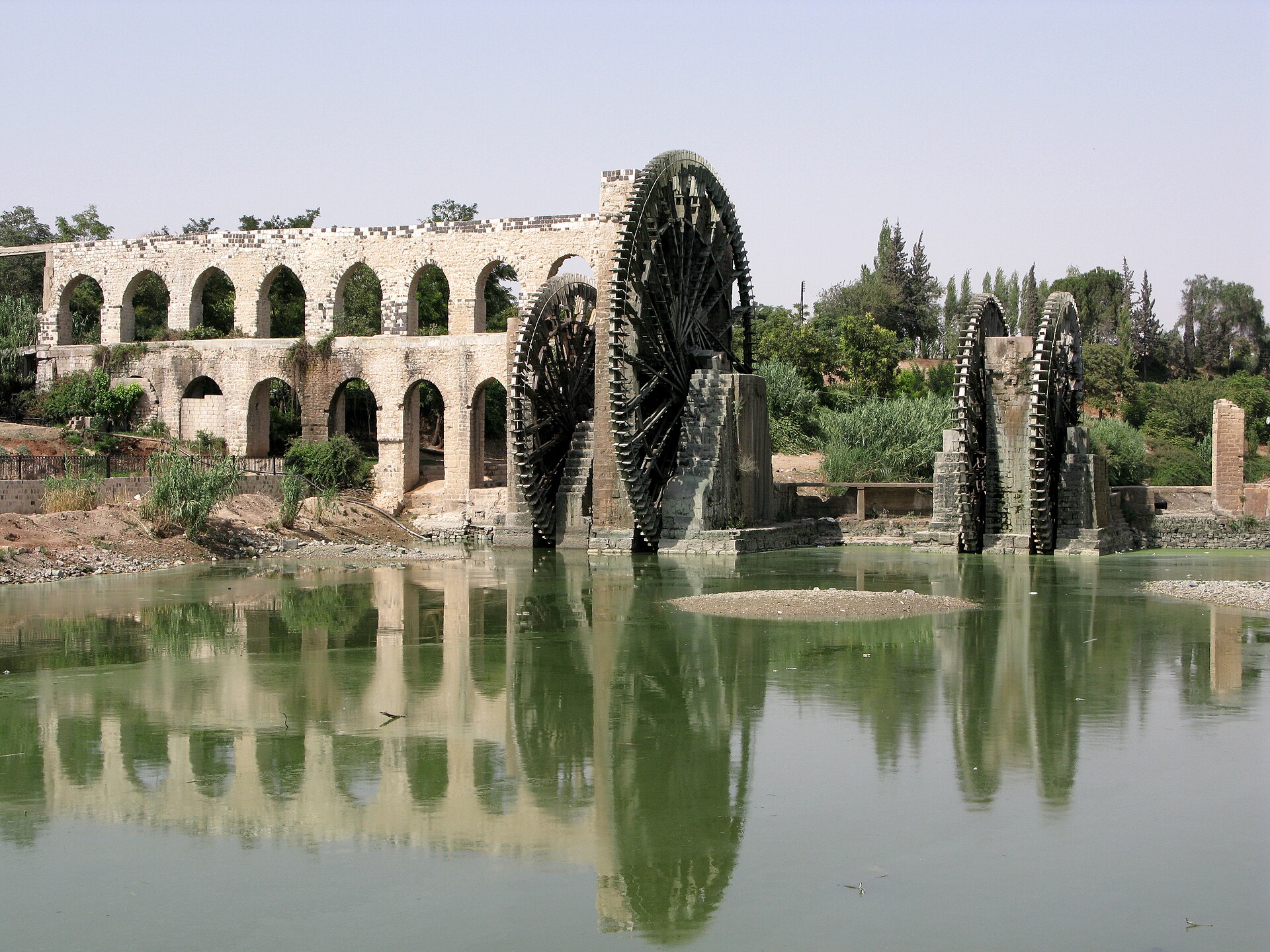 Large wooden water wheels on the Orontes river in Hama, beside a stone aqueduct.